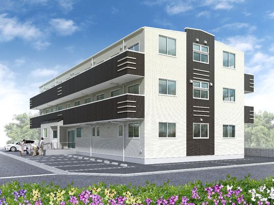 駐車場がついた３階建ての建物。玄関には階段と手すり付きのスロープがあり、敷地の周りには花が植えられている。