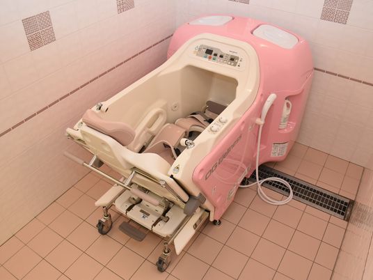 車いすの高齢女性の介助をしているピンクのポロシャツ姿の女性スタッフ。二人は屋外を散歩している。