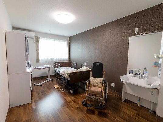 車いすが置かれたフローリングの部屋には収納家具や洗面所があり、大きな窓にはカーテンが付いている。