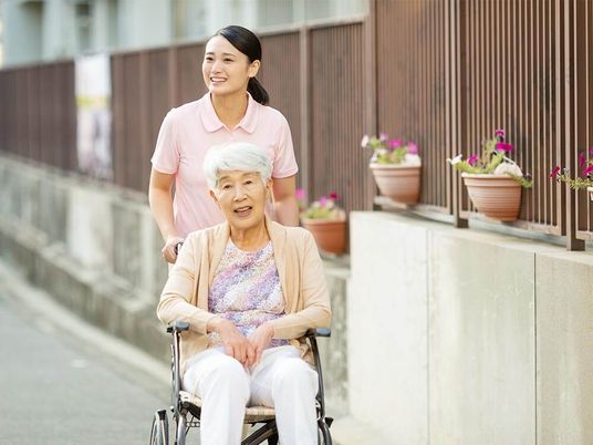 車いすの高齢女性の車いすを押しながら楽しそうな表情を浮かべ散歩をしているポロシャツを着た女性スタッフ。