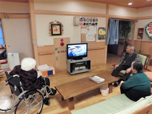 リビングでテレビを囲んでいる3名の入居者。車いす使用者もいる。