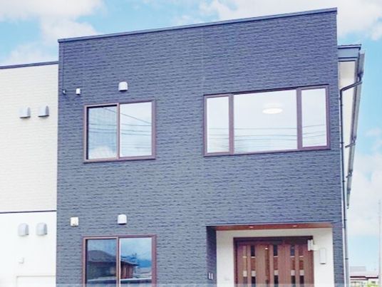 白やグレーの外壁の２階建ての建物は日当たり良好で窓がたくさん付き、玄関に屋根がある。